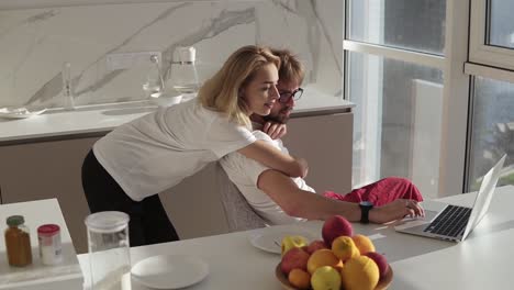 Side-view-of-bearded-man-working-on-laptop-computer-sitting-at-home-on-a-kitchen-with-panoramic-windows.-Young-wife-embracing-him-from-behind-and-inviting-for-breakfast-or-asking-to-help