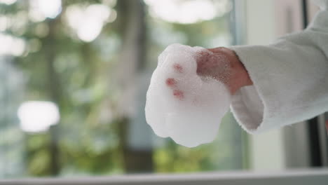 la mujer toca suavemente el baño de burbujas suave de cerca. la dama en bata de baño de terry sostiene la espuma apreciando el aroma calmante por la mañana. procedimientos de cuidado corporal en casa