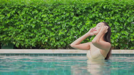 Mujer-Asiática-Sexy-Arreglándose-El-Pelo-Mojado-En-Una-Piscina-Tropical-Con-Fondo-De-Plantas-Verdes,-Cámara-Lenta-De-Cuadro-Completo