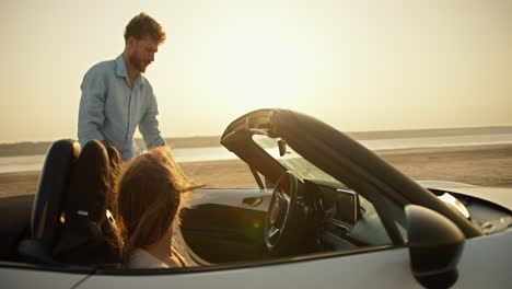 A-bearded-guy-with-curly-hair-in-a-blue-shirt-opens-the-door-of-a-white-convertible-car-and-sits-inside-next-to-his-blonde-girlfriend-against-the-river-with-a-yellow-sky-and-a-bright-sun