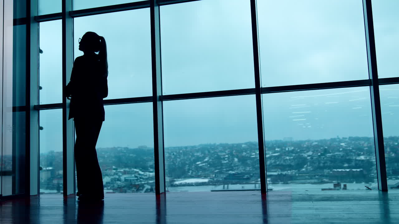 Silhouette of a woman in glasses and suit walks along the panoramic windows. City panorama at backdrop. Low angle view.