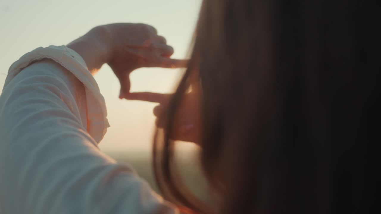 Close up rear view of woman forming rectangle shape with hands as sunlight passes through, creating glowing flare under palm during golden hour, strands of hair softly illuminated