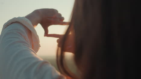 close up rear view of woman forming rectangle shape with hands as sunlight passes through, creating glowing flare under palm during golden hour, strands of hair softly illuminated