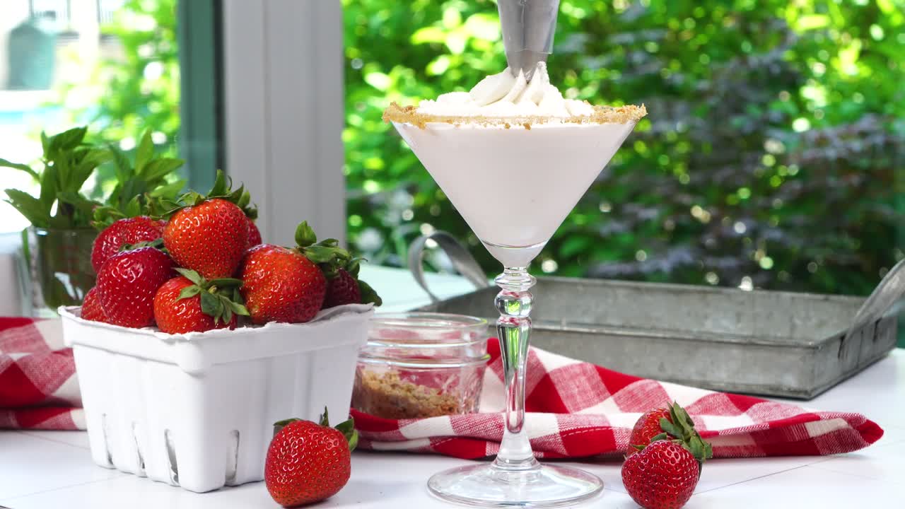 Strawberry Shortcake Martini cocktail with whipped cream, graham cracker crumbs and a fresh red strawberry. Piping the whipped cream on top. A summer time berry drink with red gingham napkin.