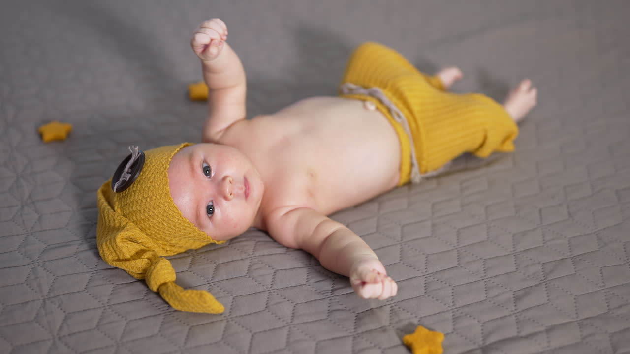 Small boy waving his arms and feet while lying on the bed. Boy in yellow costume with bare belly. Kid in a funny hat with big button.