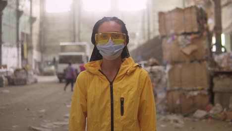 Una-Chica-Positiva-Con-Ropa-De-Trabajo-Protectora-Chaqueta-Amarilla,-Anteojos-Y-Máscara-Caminando-Por-La-Fábrica-De-Reciclaje-Y-Sonriendo