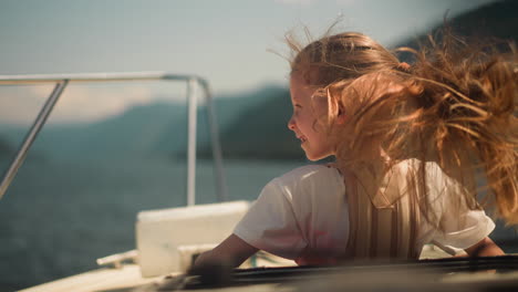 happy little girl with long hair waved by wind rides fast motorboat in tranquil sea bay. joyful child sails modern yacht in ocean on sunny day
