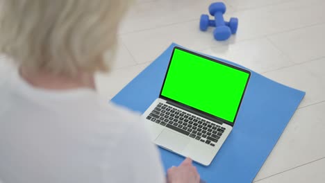 vista trasera de una anciana haciendo yoga mientras mira una computadora portátil con pantalla de tecla cromada