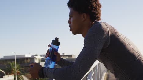 fit african american man exercising in city, resting on footbridge, drinking from water bottle