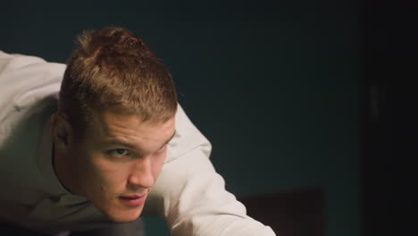 close-up of young man in white shirt leaning over green pool table, gripping cue stick with intense focus. dark background enhances concentration and tension as he prepares for precise shot