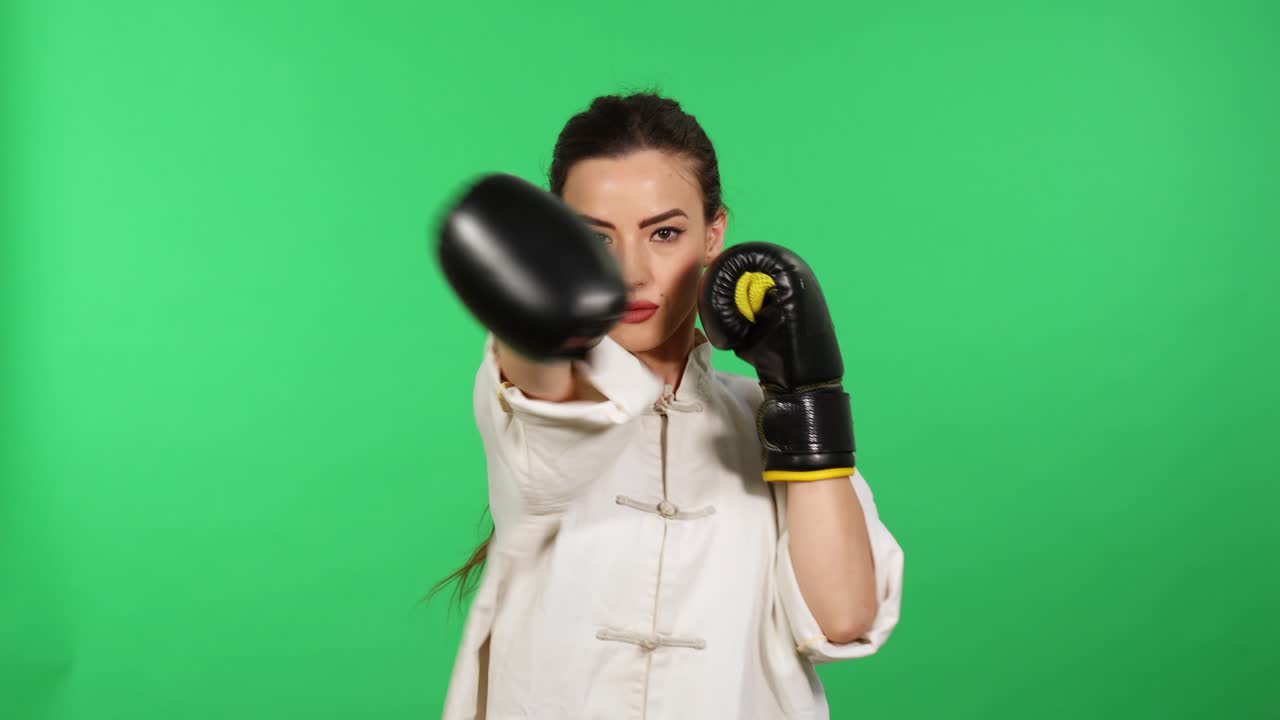 joven morenita mujer con guantes de boxeo y blanco kung fu wushu unifrom puñetazos hacia la cámara con pantalla verde en segundo plano.