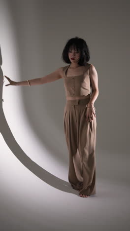 vertical video: model posing in stylish beige outfit, leaning against wall in fashion studio