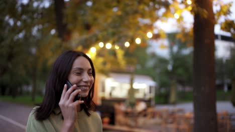 Portrait-of-a-brunette-in-a-green-sweater,-who-speaks-by-phone-against-the-backdrop-of-the-bright-lights-of-a-cafe-in-the-park