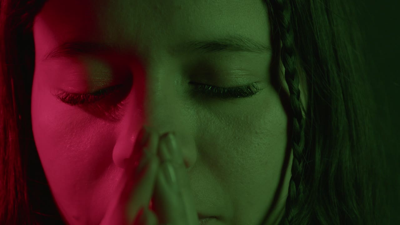 A close-up shot captures a girl praying, her forehead and braided hair partially visible, enveloped in low, moody green lighting, conveying a sense of peace and introspection
