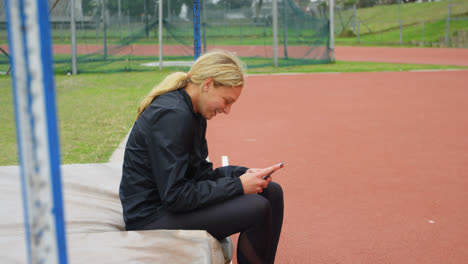 Vista-Lateral-De-Una-Atleta-Caucásica-Usando-Un-Teléfono-Móvil-En-Un-Recinto-Deportivo-4k