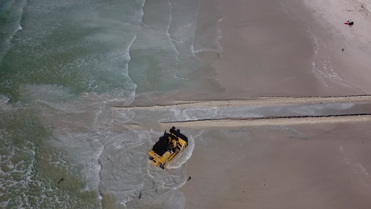 A heavy machinery Caterpillar bulldozer opens a passage for water to flow from Zandvlei Estuary Nature Reserve into the Muizenberg beach. Static aerial view of the bulldozer reversing out of the sea.
