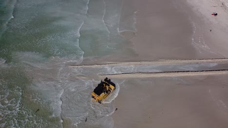 a heavy machinery caterpillar bulldozer opens a passage for water to flow from zandvlei estuary nature reserve into the muizenberg beach. static aerial view of the bulldozer reversing out of the sea.