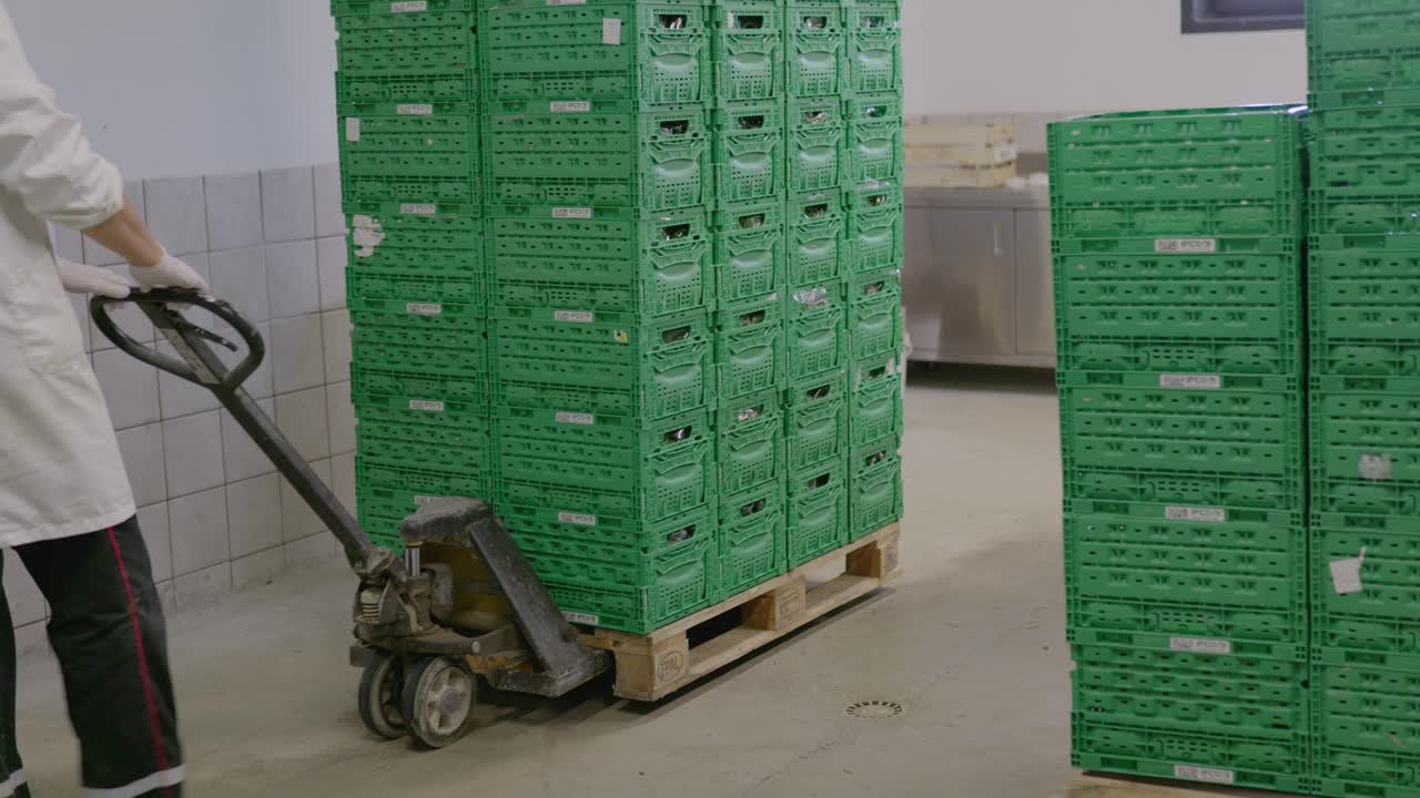 Worker using a pallet jack to move a tall stack of green crates in an industrial facility