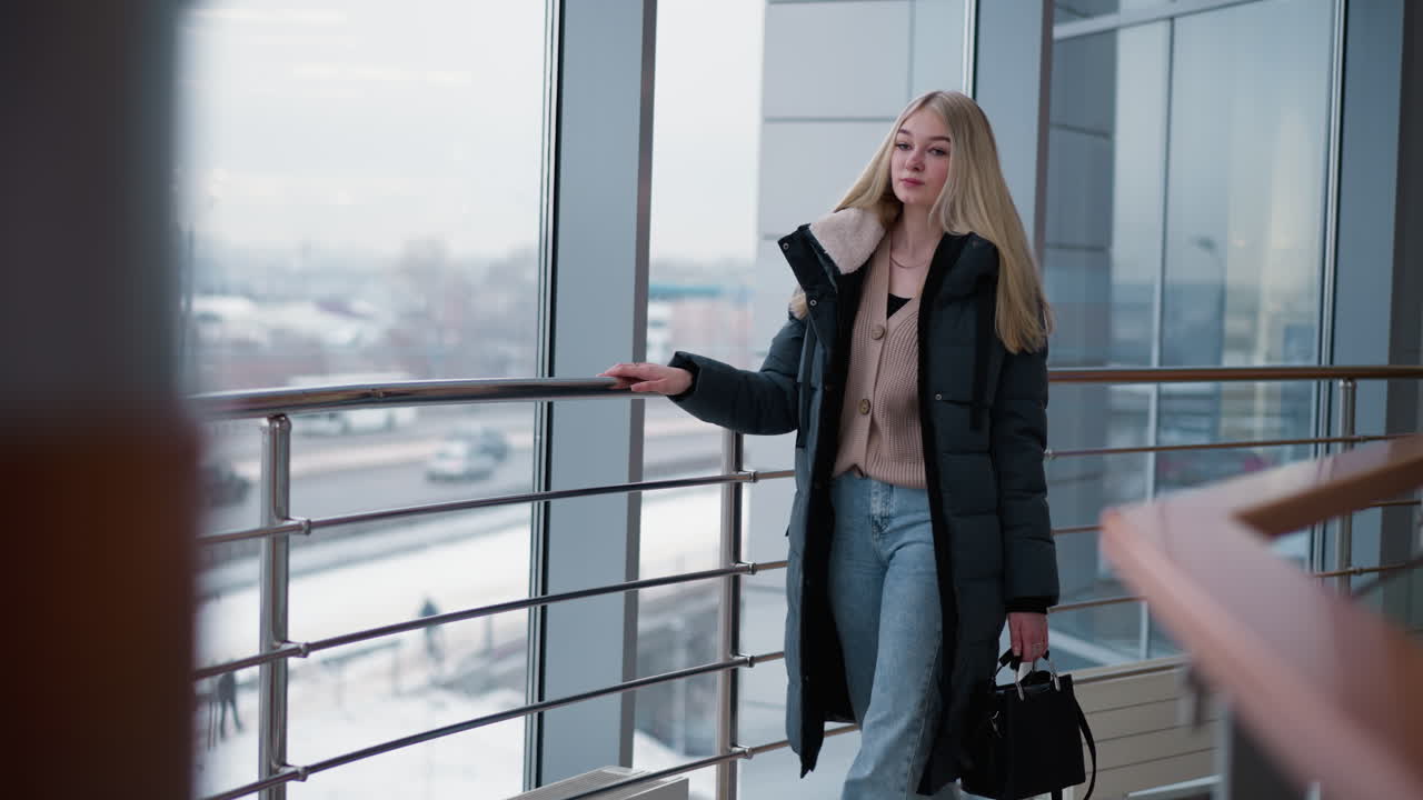 Elegant lady walking near iron railing, gliding hand along metal rail, gazing at city view through large glass windows in modern building, dressed in stylish winter outfit with long blonde hair