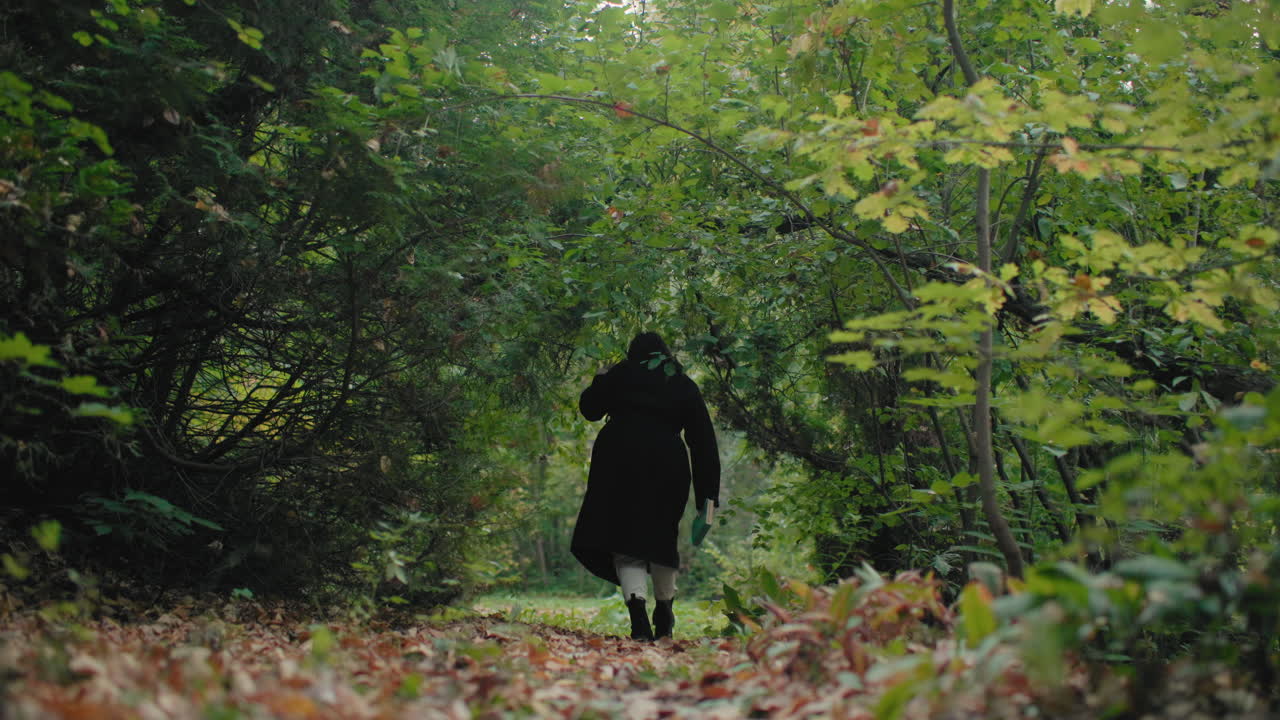 Back view of wandering lady in black coat walking into forest with book in hand, surrounded by green leaves and soft light, slow solitary stroll toward deeper woods, calm autumn mood
