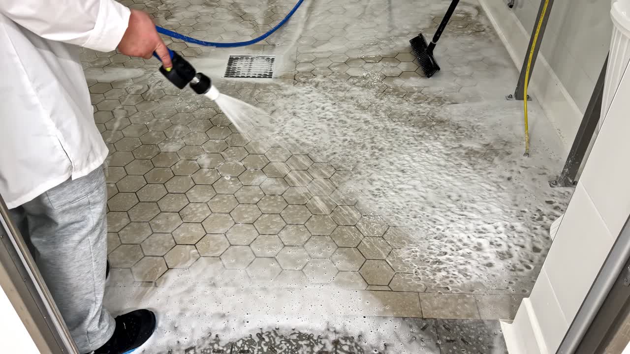 Worker rinsing foam and cleaning solution from tiled flooring inside a food-grade facility, directing water toward the drain during sanitation procedures