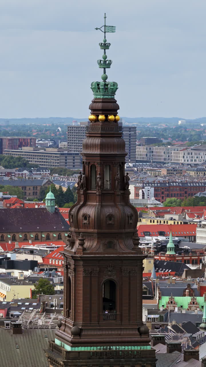 Aerial drone view of the ornate spire of Christiansborg Palace, crowned with golden spheres and intricate details, overlooking the cityscape of Copenhagen, Denmark. Vertical