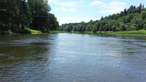 aerial: slow and low flying above the river with forest on each side on a sunny day with sky and clouds visible in the background