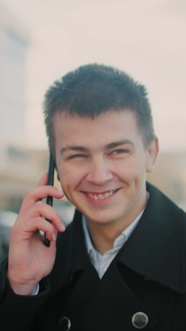 흑색 코트를 입은 남자가 웃으며 전화로 대화하면서 현대적인 유리 건물, 주차된 자동차, 부드러운 빛, 그리고 즐거운 분위기를 특징으로하는 활기찬 도시 지역에서 흥분합니다.