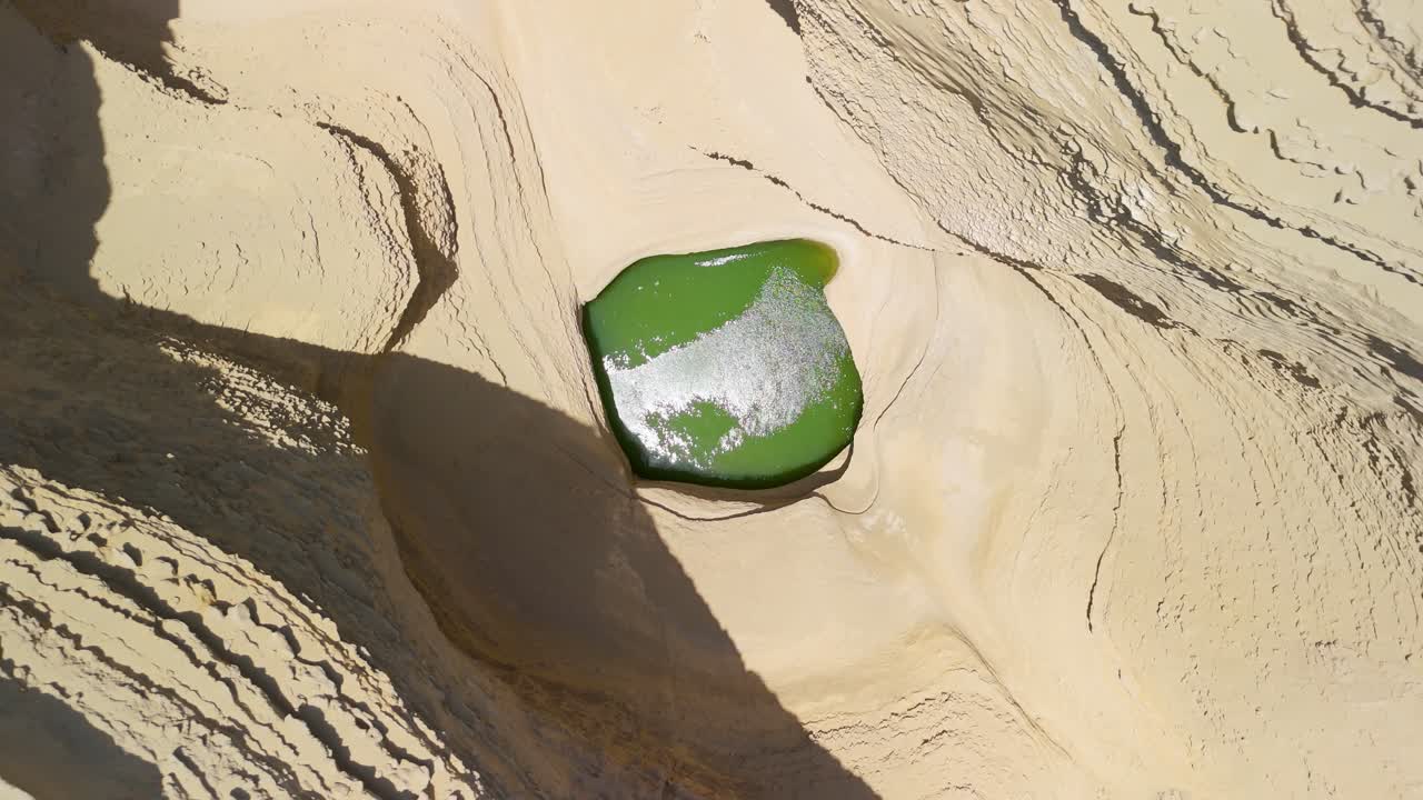 Spinning top-down aerial rise above strikingly green water pool nestled within dry, layered geological formations of Cañon de los Perdidos or Canyon of the Lost in the Ocucaje desert, Ica Peru