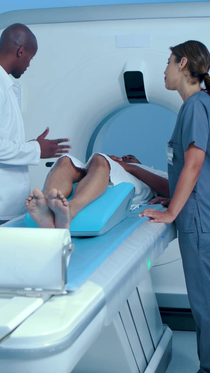 Video verticale: Medico che arriva in camice usando un tablet in sala TC, infermiera in divisa che tiene un uomo su un cuscino a cuneo