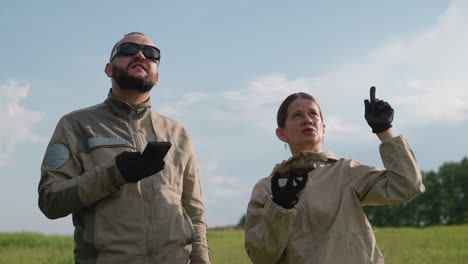 man and woman colleagues focus on devices while discussing in grassy field under blue sky, woman gesturing to shield sunlight as they collaborate on mobile research outdoors