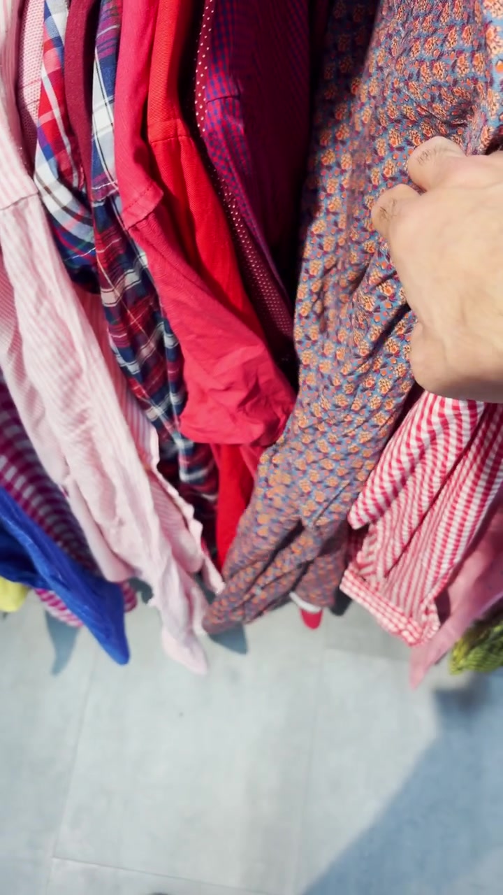 Point of view vertical UGC style reel of person looking for vintage clothes on racks in retro sustainable zero waste store. Hipster in thrift shop or flea market. Browsing used wear on hangers on sale
