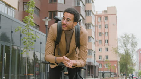 young american man in formal clothes and backpack looking at the camera and around while leaning on bicycle handlebars in the street