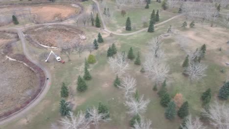 los ojos de los pájaros aéreos sobrevuelan los estanques secos hechos por el hombre, el centro comunitario, la poda de árboles machinary en un parque en una tarde completa con pinos dispersos y arces desnudos1-3