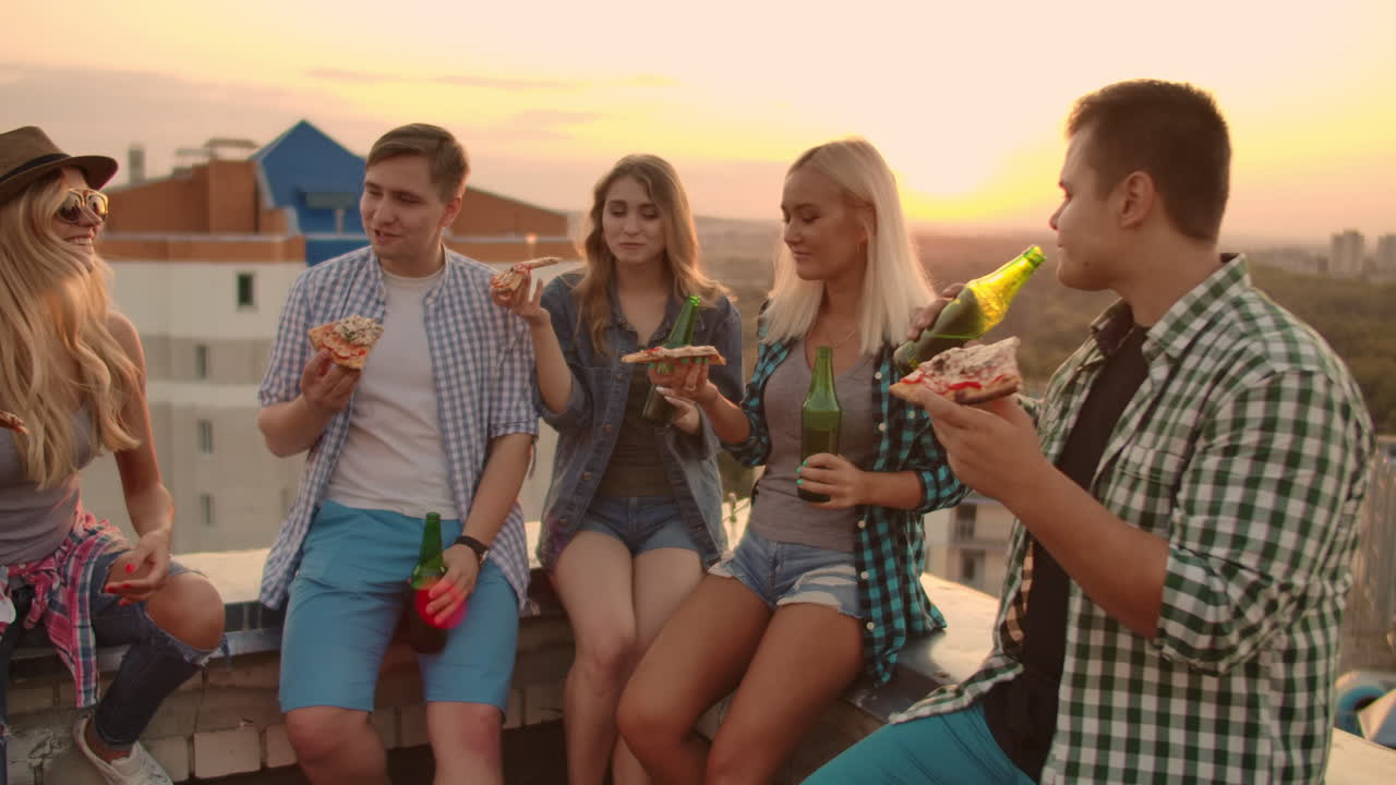 Young frieds at sunset on the roof enjoy pizza and beer. They sit and talk to each other.