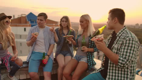 young frieds at sunset on the roof enjoy pizza and beer. they sit and talk to each other.