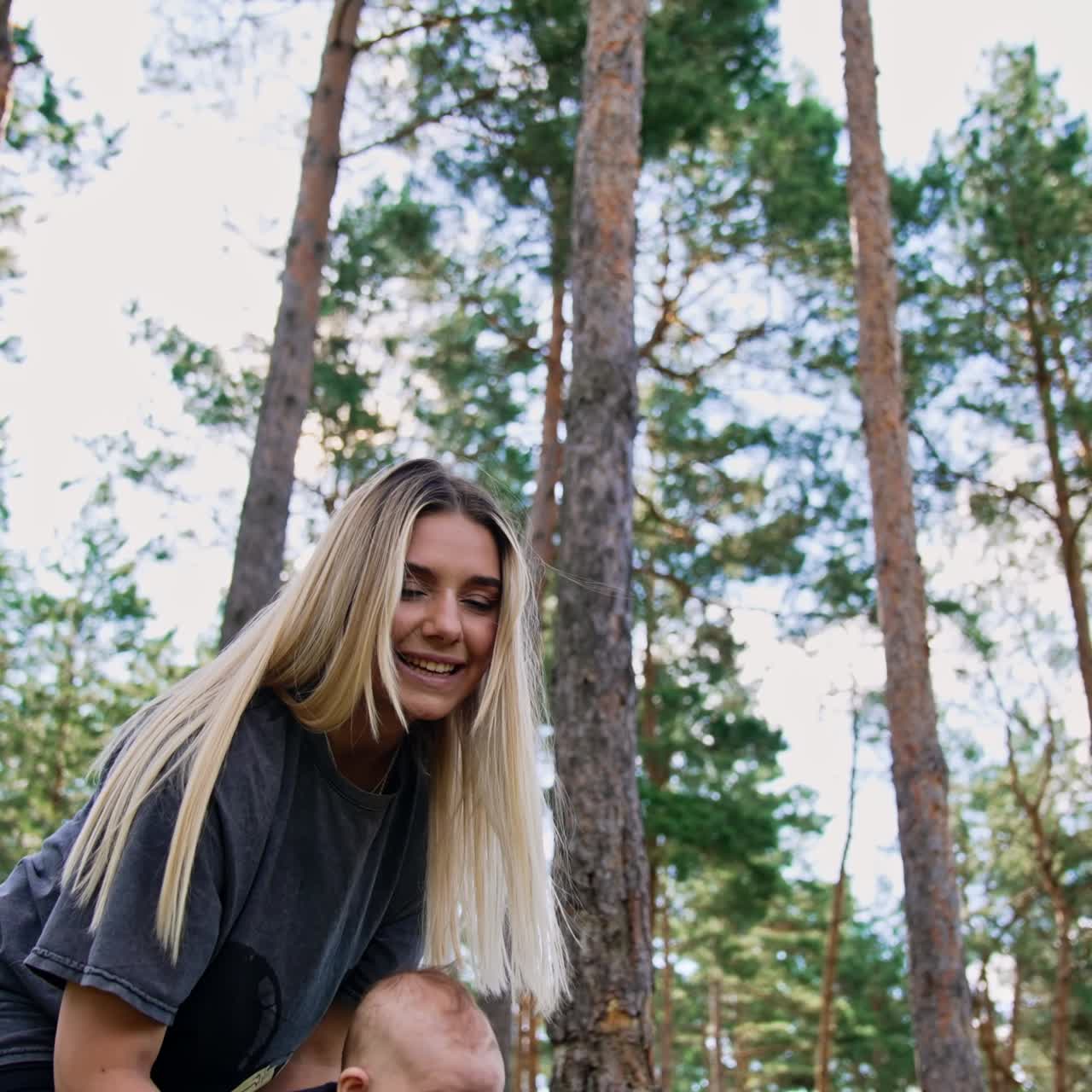 Pretty blonde woman holding her child in hands. Caring mom waving a baby in front of her. Low angle view. Nature backdrop