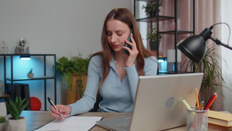 Young-woman-freelancer-answering-to-client-on-mobile-phone-call,-using-laptop-computer,-home-office