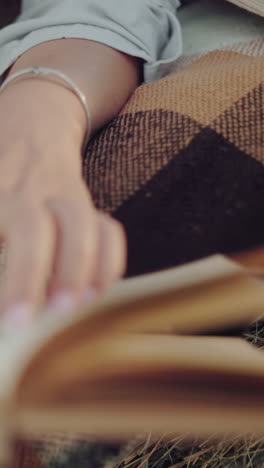 primer plano de una mano de persona con un brazalete, hojeando páginas de libros mientras se acuesta en una manta a cuadros sobre una bala de heno, evocando un entorno de lectura rústico y pacífico
