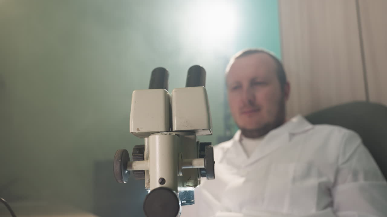 una vista de cerca de un científico examinando intensamente muestras a través de un microscopio en un laboratorio, luego se mueve hacia atrás desde el microscopio, el científico está en foco, con una bata blanca de laboratorio