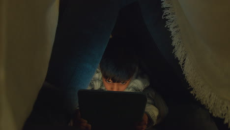 primer plano de un niño en el campamento hecho en casa hecho de cojines jugando con la tableta digital por la noche