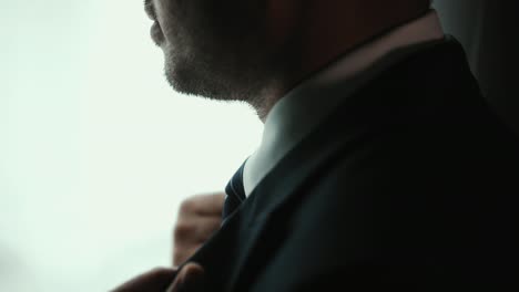 confident businessman in classic blue suit tying or adjust necktie near window in hotel room at the morning. handsome man wearing a nice suit on wedding day.