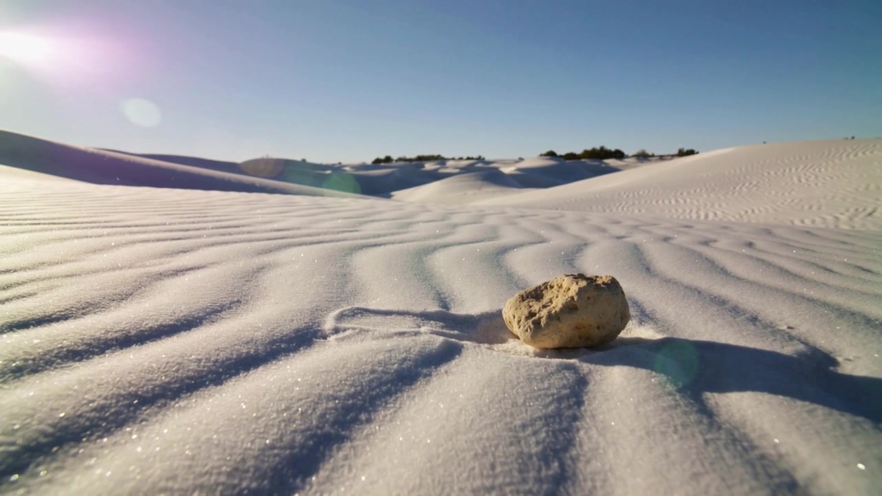 Câmera em movimento de baixo ângulo mostrando rocha em areia ondulada em um campo de dunas, com o sol a brilhar destacando a textura
