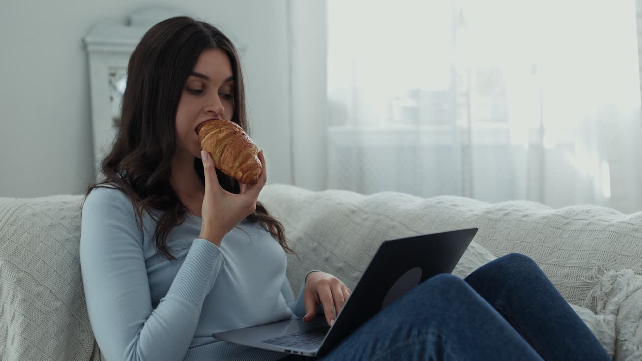 mujer comiendo croissant y trabajando en la computadora portátil en casa