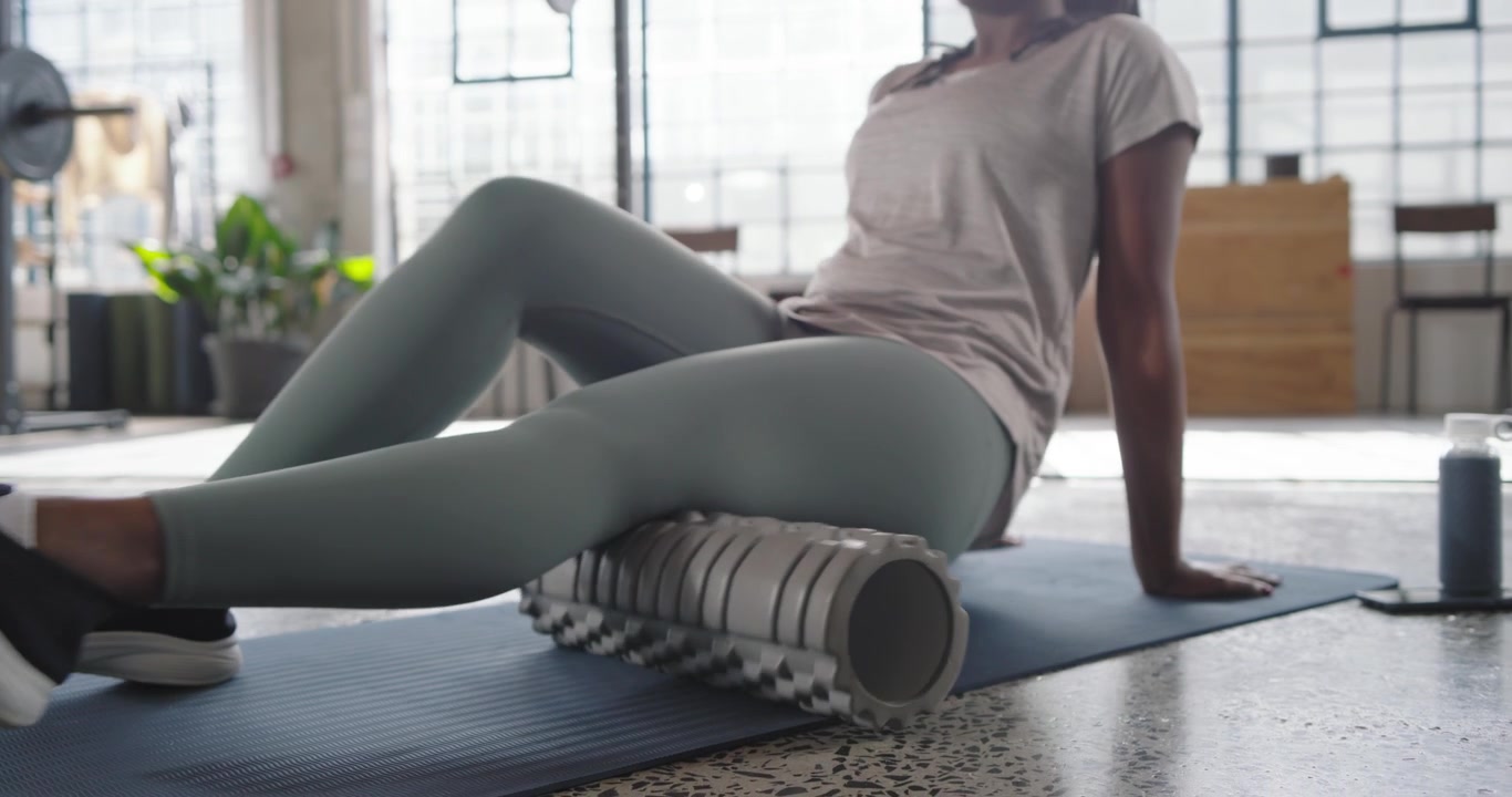 African woman in sage leggings placing foam-roller under right glute on blue mat rolling for relief