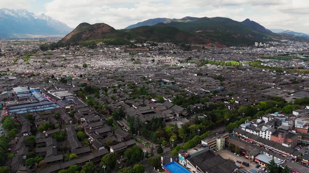 Lijiang Old Town buildings extend toward the horizon with distant hills under blue sky, panoramic aerial establishing overview