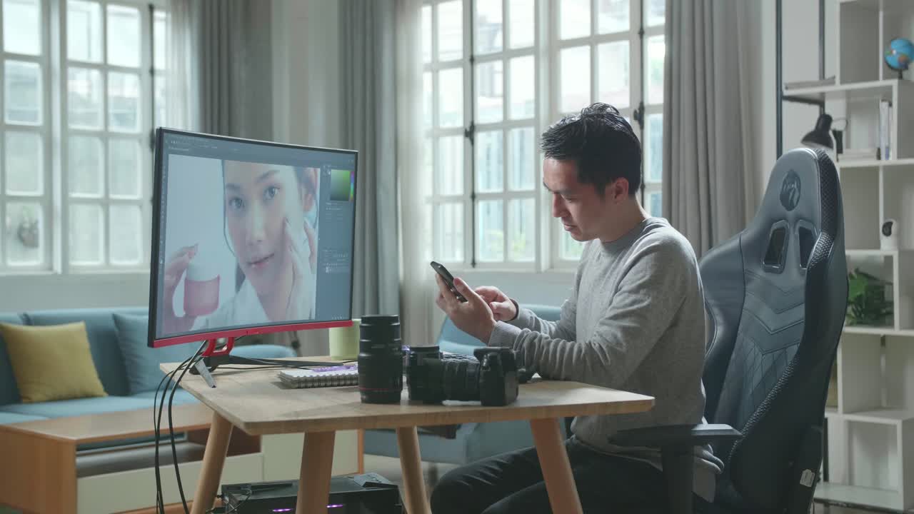 camarógrafo asiático en camiseta de mangas largas y pantalones negros usando teléfono móvil mientras usa computadora de escritorio para editar fotos en casa.