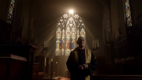 a clergyman stands in a dimly lit church, illuminated by light streaming through a stained glass window