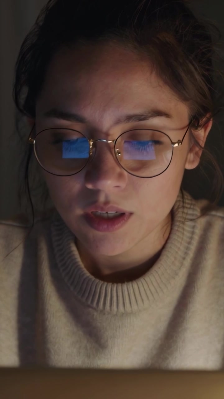 Video verticale: Donna in maglione che legge a casa al laptop sotto la luce dello schermo, sistemandosi gli occhiali rotondi.