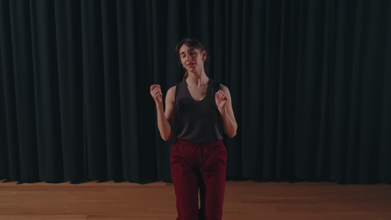 Kvindelig moderne danser, der udfører ekspressiv solo-bevægelse med åbne arme på en teaterscene med mørkt gardinbagtæppe og varmt trægulv